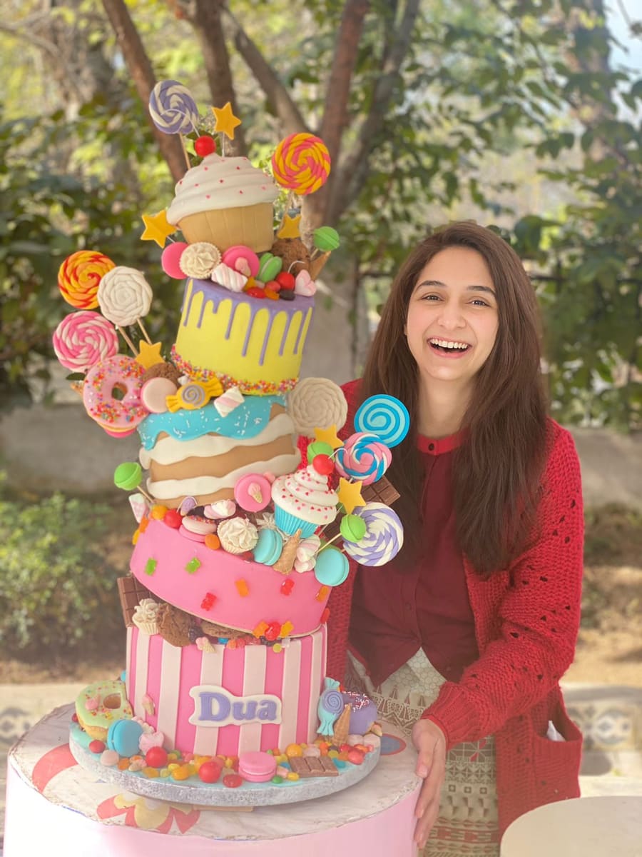 The founder of Cake Me Away smiling beside a towering topsy turvy birthday cake covered in lollipops, sweets and a giant cupcake.